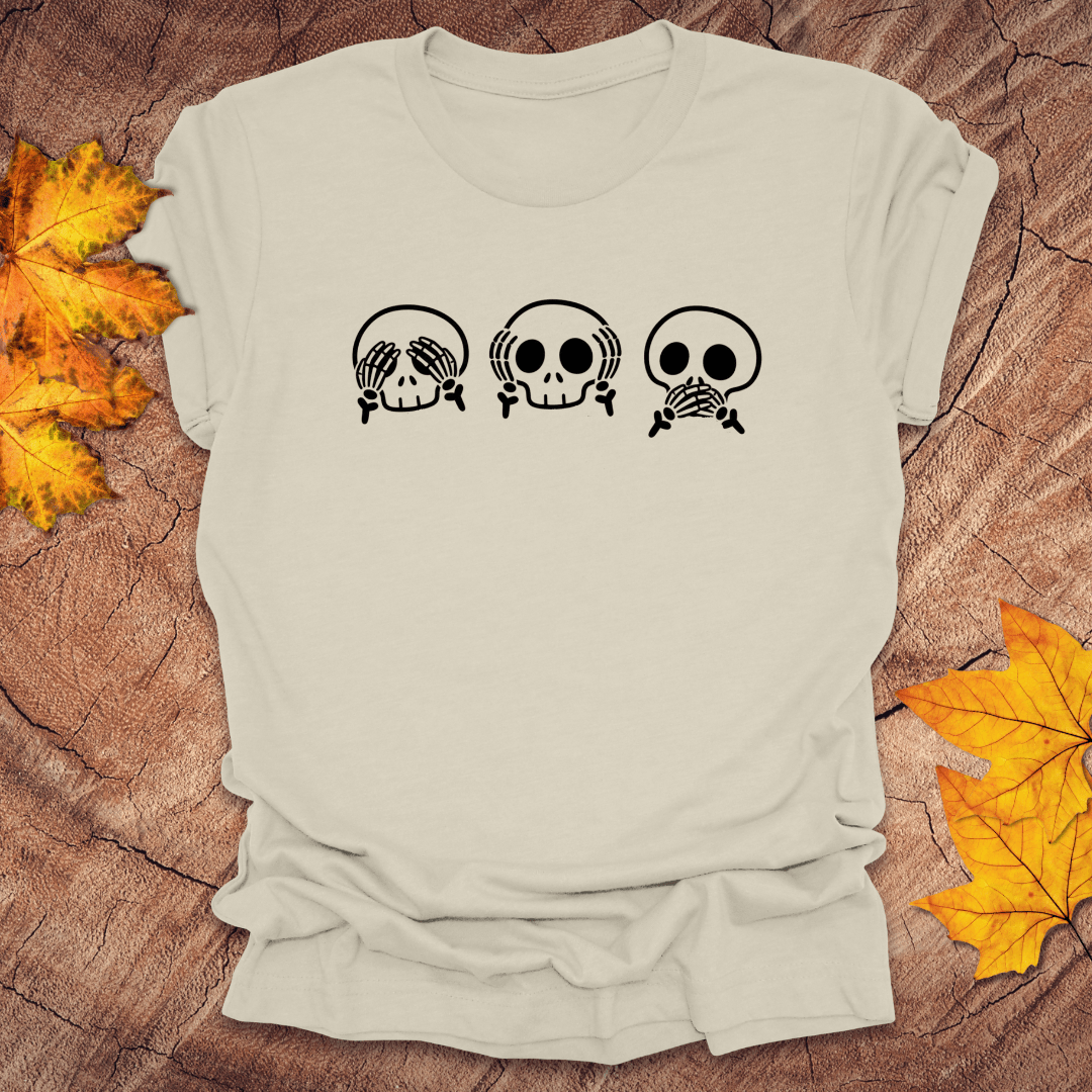 Beige tshirt with 3 skulls simulating see no evil, hear no evil, speak no evil, on a wood background with fall leaves.