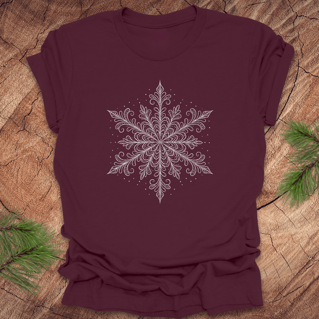 Maroon t-shirt with a white snowflake design on a wooden surface with pine branches.
