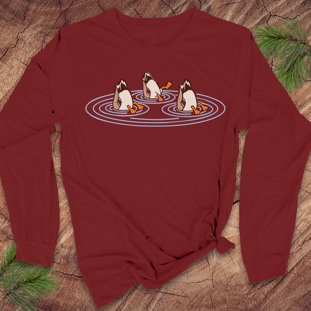 Cardinal red long-sleeve t-shirt with three Mallards dabbling bottoms-up in rippling pond water, laid flat on a rustic wood background with pine branches.