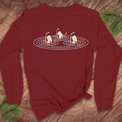 Cardinal red long-sleeve t-shirt with three Mallards dabbling bottoms-up in rippling pond water, laid flat on a rustic wood background with pine branches.