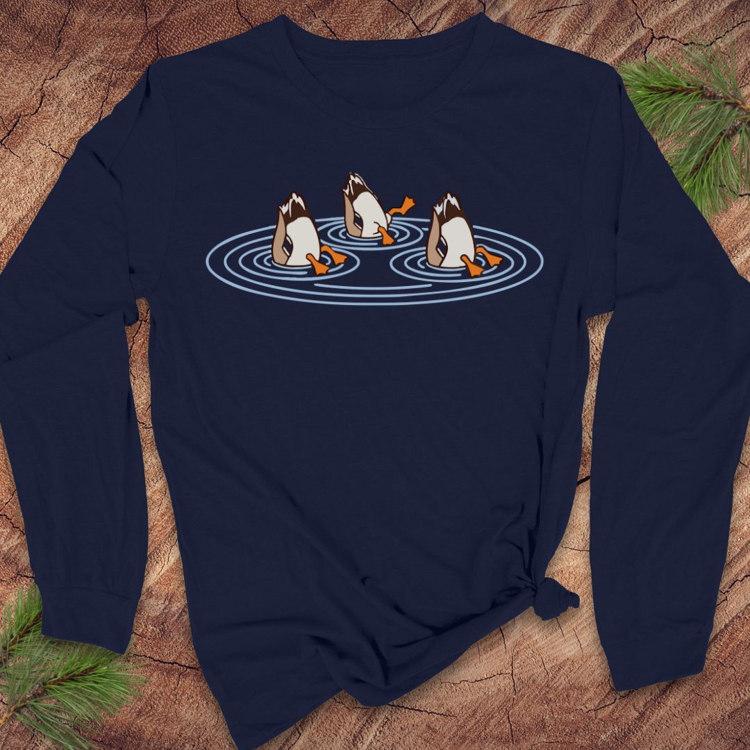 Navy long-sleeve t-shirt with three Mallards dabbling bottoms-up in rippling pond water, laid flat on a rustic wood background with pine branches.