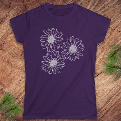 A purple short-sleeve crewneck t-shirt featuring a white line art graphic of three daisy flowers on the front, displayed flat on a textured wood background with pine branches in the corners.
