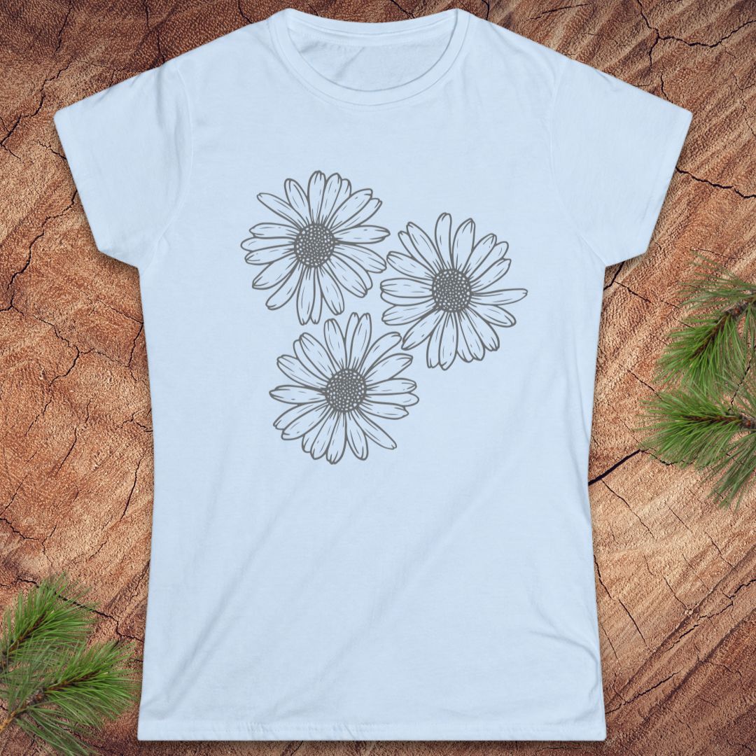 A light blue short-sleeve crewneck t-shirt featuring a black line art graphic of three daisy flowers on the front, displayed flat on a rustic wood background with pine branches in the corners.
