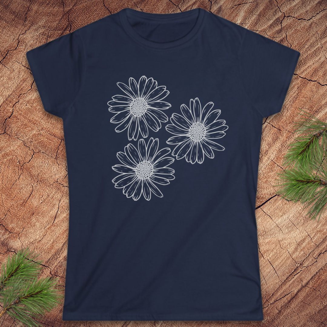 A navy blue short-sleeve crewneck t-shirt featuring a white line art graphic of three daisy-like flowers on the front, displayed flat on a rustic wood background with pine needles in the corners.