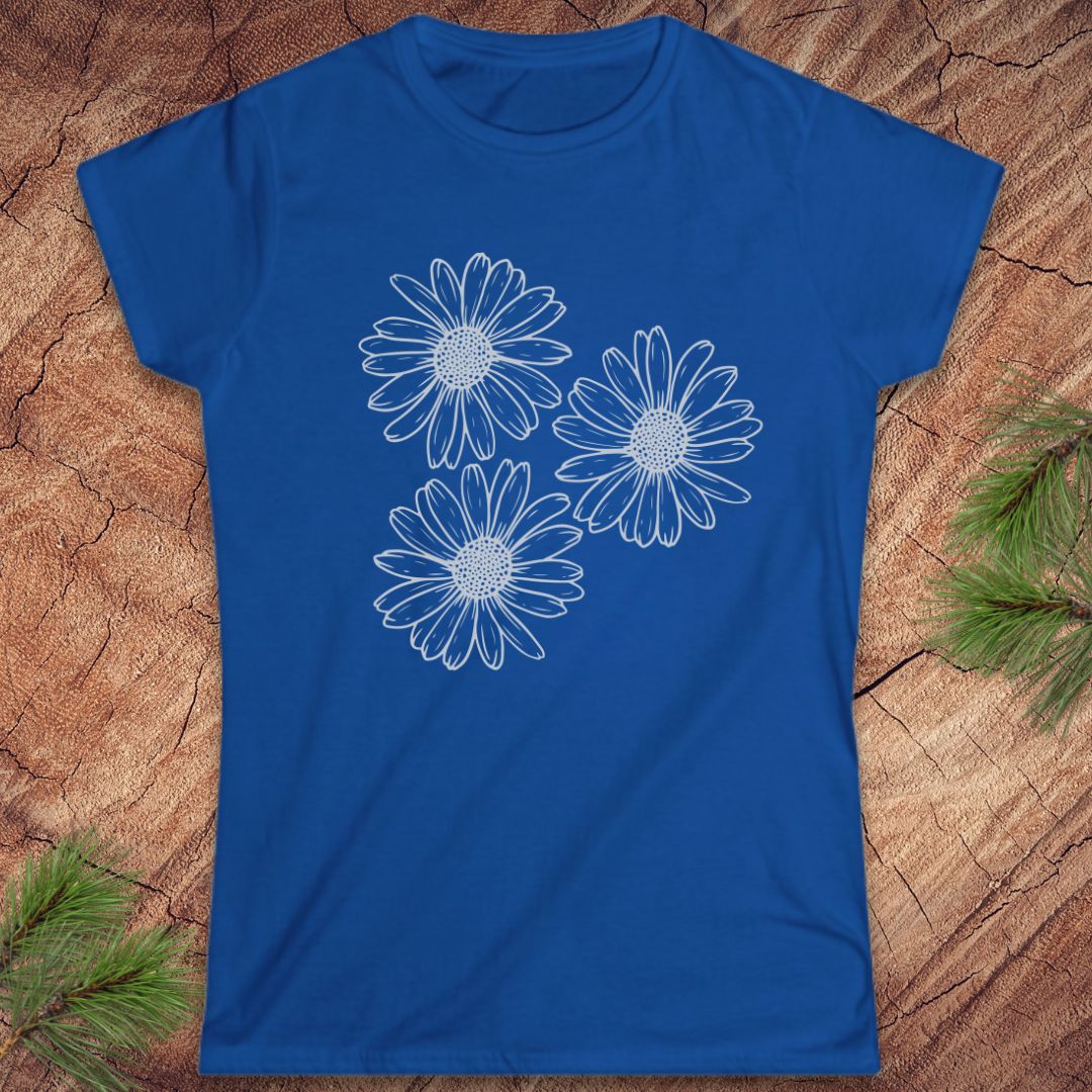 A royal blue short-sleeve t-shirt with a white line-art graphic of three daisy-like flowers on the front, laid flat on a textured wood background with pine branches in the corners.