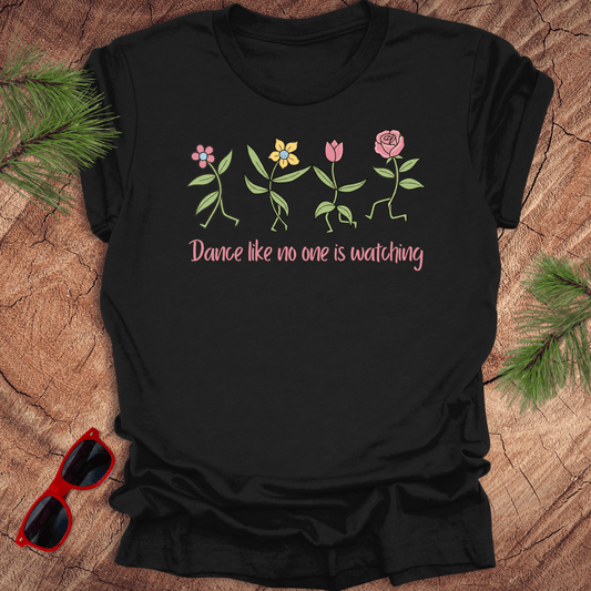 Black t-shirt with flower dancing design and text, 'Dance like no one is watching' on a wooden surface with sunglasses and greenery.