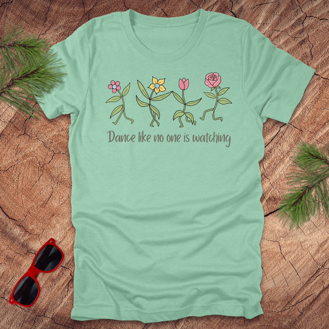 Mint green t-shirt with flower dancing design and text, 'Dance like no one is watching' on a wooden surface with sunglasses and greenery.