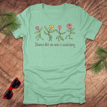 Mint green t-shirt with flower dancing design and text, 'Dance like no one is watching' on a wooden surface with sunglasses and greenery.