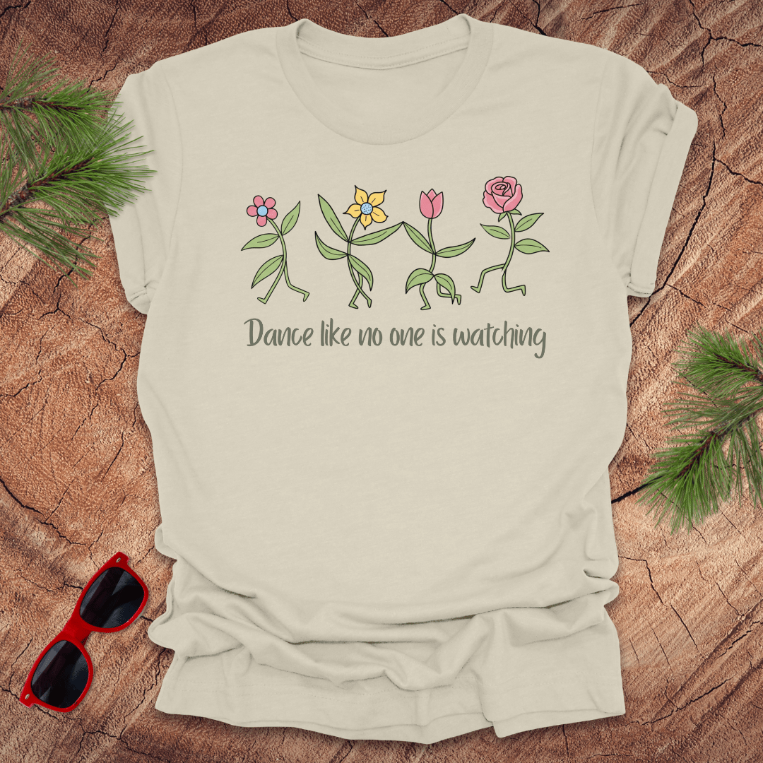 Beige t-shirt with flower dancing design and text, 'Dance like no one is watching' on a wooden surface with sunglasses and greenery.