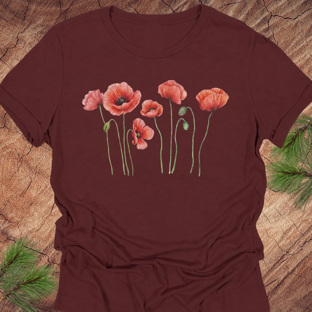 Maroon women's relaxed t-shirt with watercolor red-orange poppies on slender green stems, laid flat on a rustic wood background with pine branches.