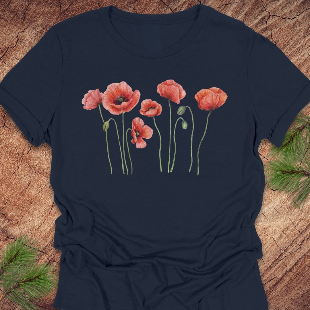 Navy women's relaxed t-shirt with watercolor red-orange poppies on slender green stems, laid flat on a rustic wood background with pine branches.