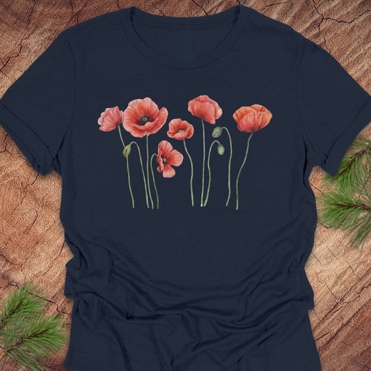 Navy women's relaxed t-shirt with watercolor red-orange poppies on slender green stems, laid flat on a rustic wood background with pine branches.