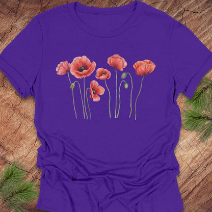 Purple women's relaxed t-shirt with watercolor red-orange poppies on slender green stems, laid flat on a rustic wood background with pine branches.