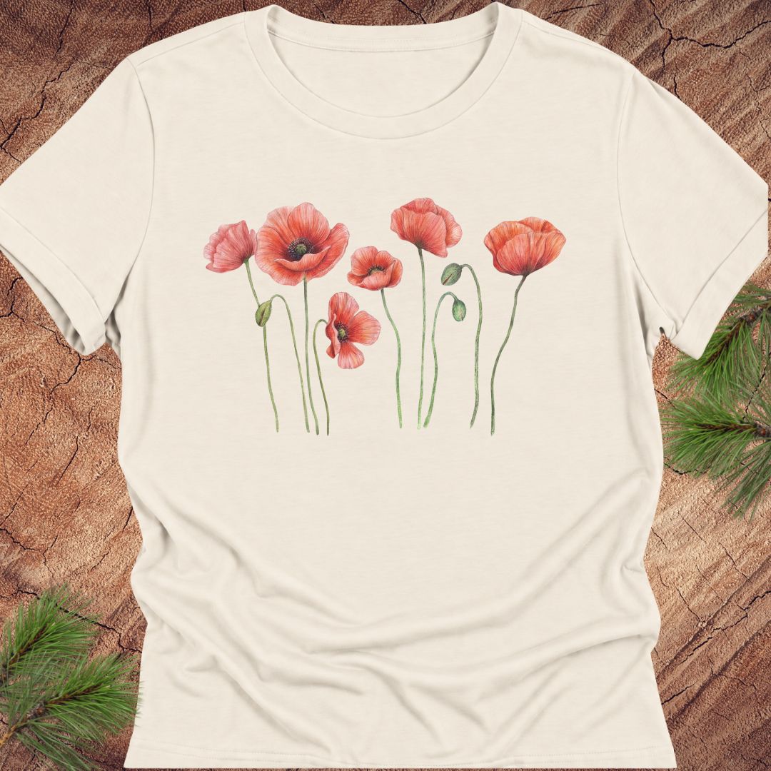 Natural cream women's relaxed t-shirt with watercolor red-orange poppies on slender green stems, laid flat on a rustic wood background with pine branches.