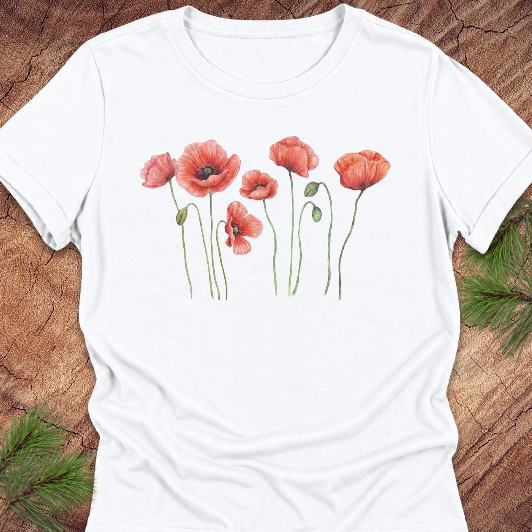 White women's relaxed t-shirt with watercolor red-orange poppies on slender green stems, laid flat on a rustic wood background with pine branches.