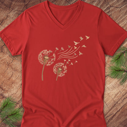 A red V-neck t-shirt laid flat on a rustic wood background with pine branches in the corners. The shirt features a graphic print of two dandelions blowing seed heads into the wind, with small birds flying away.