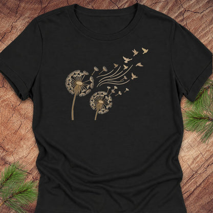 A black short-sleeve crewneck t-shirt featuring a gold graphic of two dandelions with seeds blowing away as wind, accompanied by small birds flying to the right. The shirt is displayed flat on a rustic wood background with pine needles in the corners.