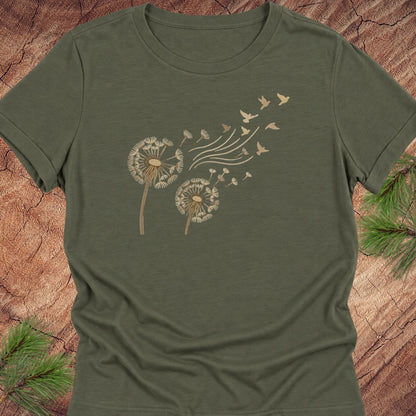 An olive green short-sleeve t-shirt with a graphic of two dandelions and birds blowing seeds, displayed flat on a rustic wood background with pine branches.