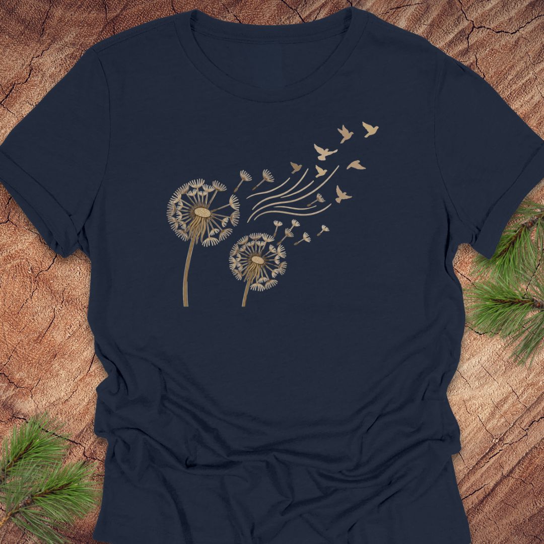 A navy blue short-sleeve t-shirt with a gold graphic of two dandelions blowing seeds into the wind, accompanied by small flying birds. The shirt is laid flat on a rustic wood background with pine needles in the corners.