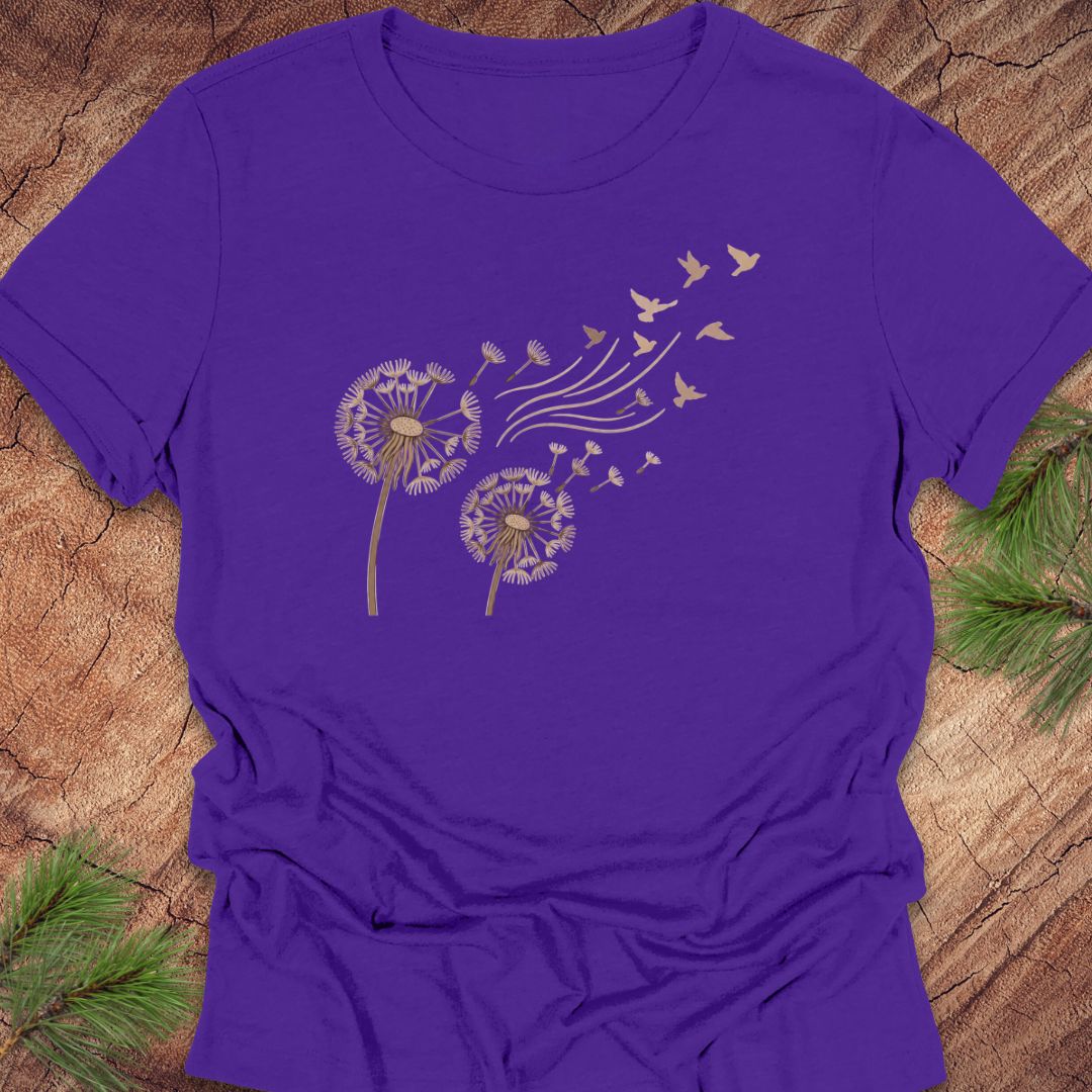 A purple short-sleeve crewneck t-shirt with a graphic of two dandelions and birds in gold and white, laid flat on a rustic wood background with pine branches.