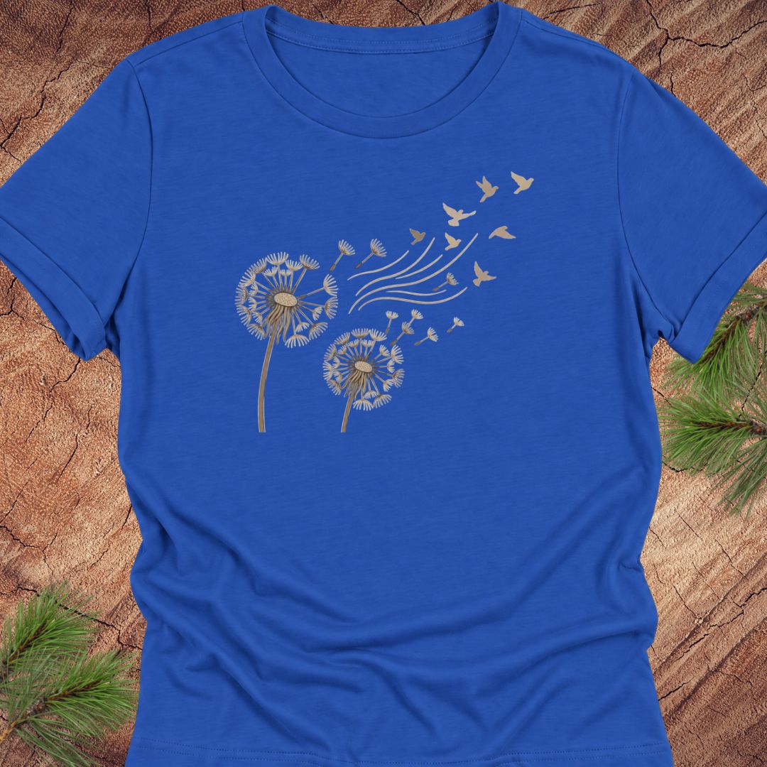 A royal blue short-sleeve crewneck t-shirt featuring a graphic of two dandelions with seeds blowing away as birds, laid flat on a rustic wood background with pine branches.