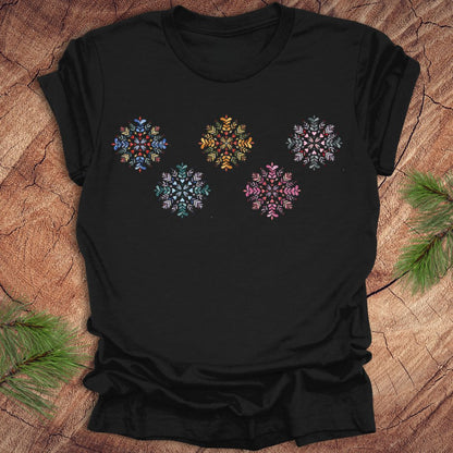 Black t-shirt with colorful folk art snowflake designs on a wooden surface with pine branches.