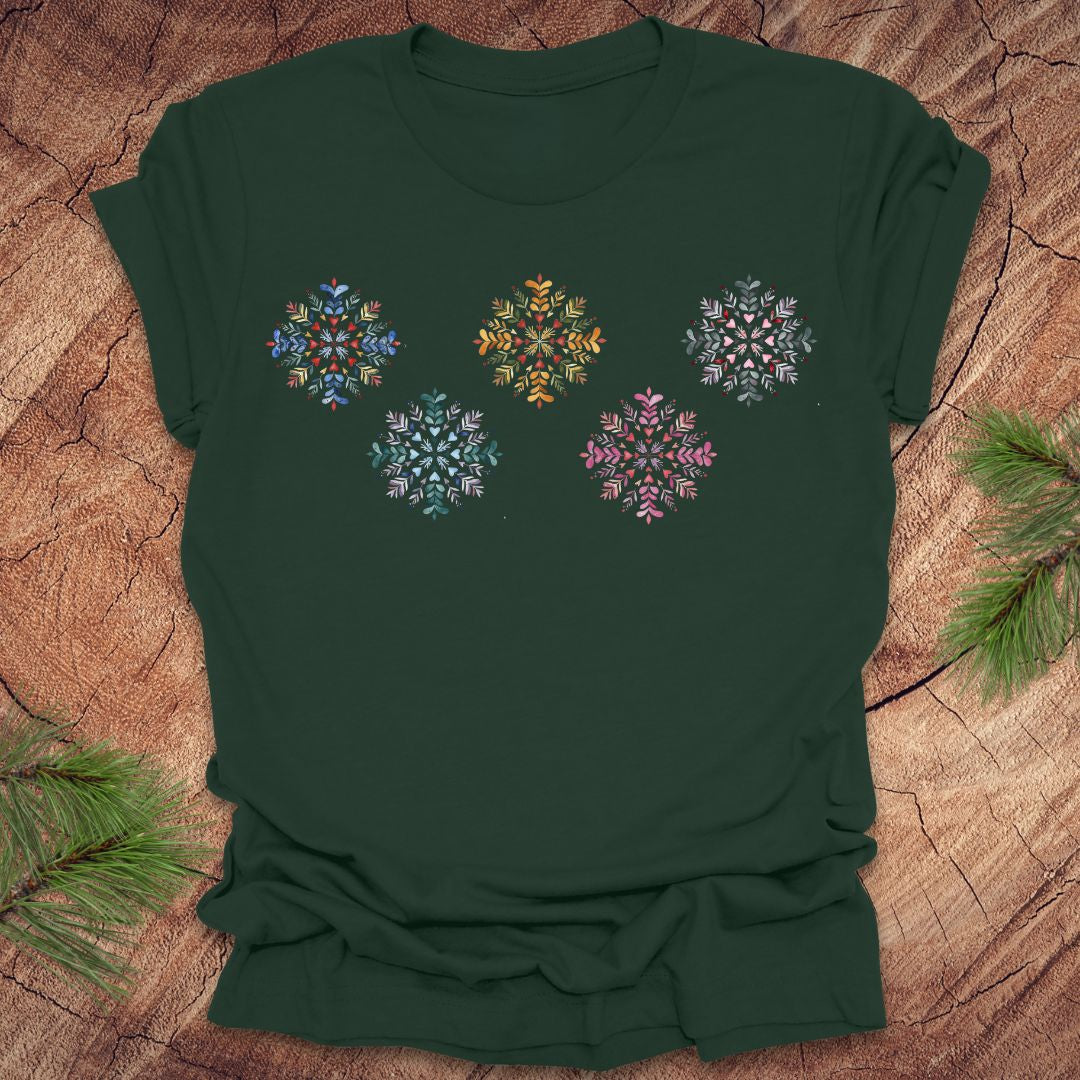 Green t-shirt with colorful folk art snowflake designs on a wooden surface with pine branches.
