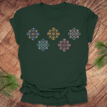 Green t-shirt with colorful folk art snowflake designs on a wooden surface with pine branches.
