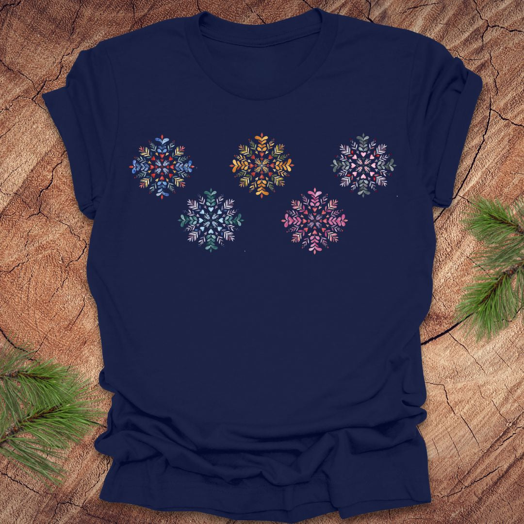 Navy blue t-shirt with colorful folk art snowflake designs on a wooden surface with pine branches.