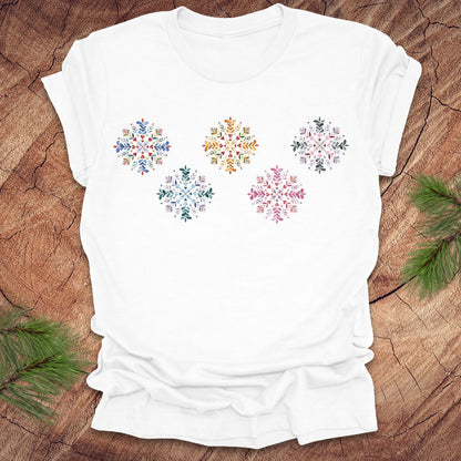 White t-shirt with colorful folk art snowflake designs on a wooden surface with pine branches.