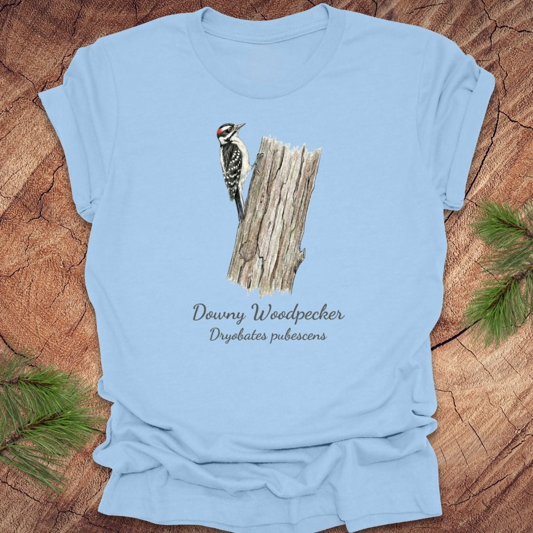 Baby blue t-shirt featuring a downy woodpecker perched on a tree with text, on a wood background.