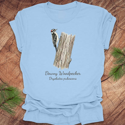 Baby blue t-shirt featuring a downy woodpecker perched on a tree with text, on a wood background.