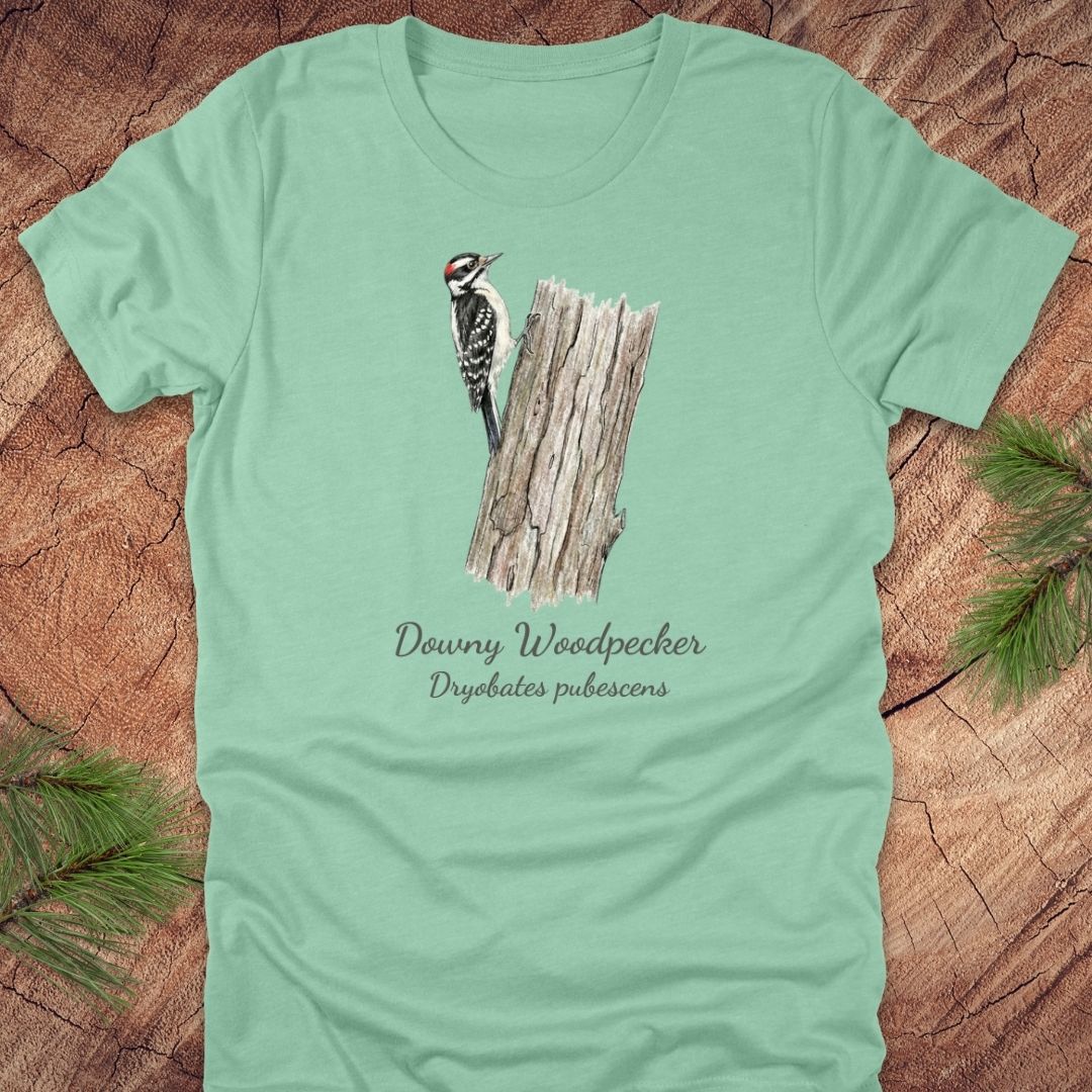 Mint green t-shirt featuring a downy woodpecker perched on a tree with text, on a wood background.
