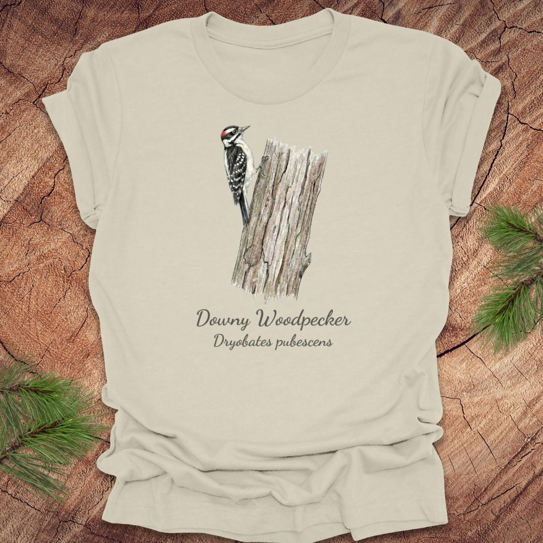 Beige t-shirt featuring a downy woodpecker perched on a tree with text, on a wood background.