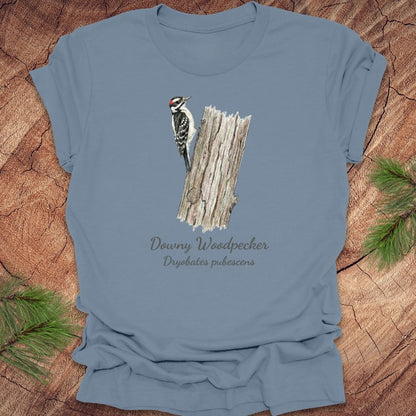 Steel blue t-shirt featuring a downy woodpecker perched on a tree with text, on a wood background.