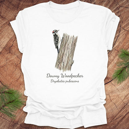 White t-shirt featuring a downy woodpecker perched on a tree with text, on a wood background.