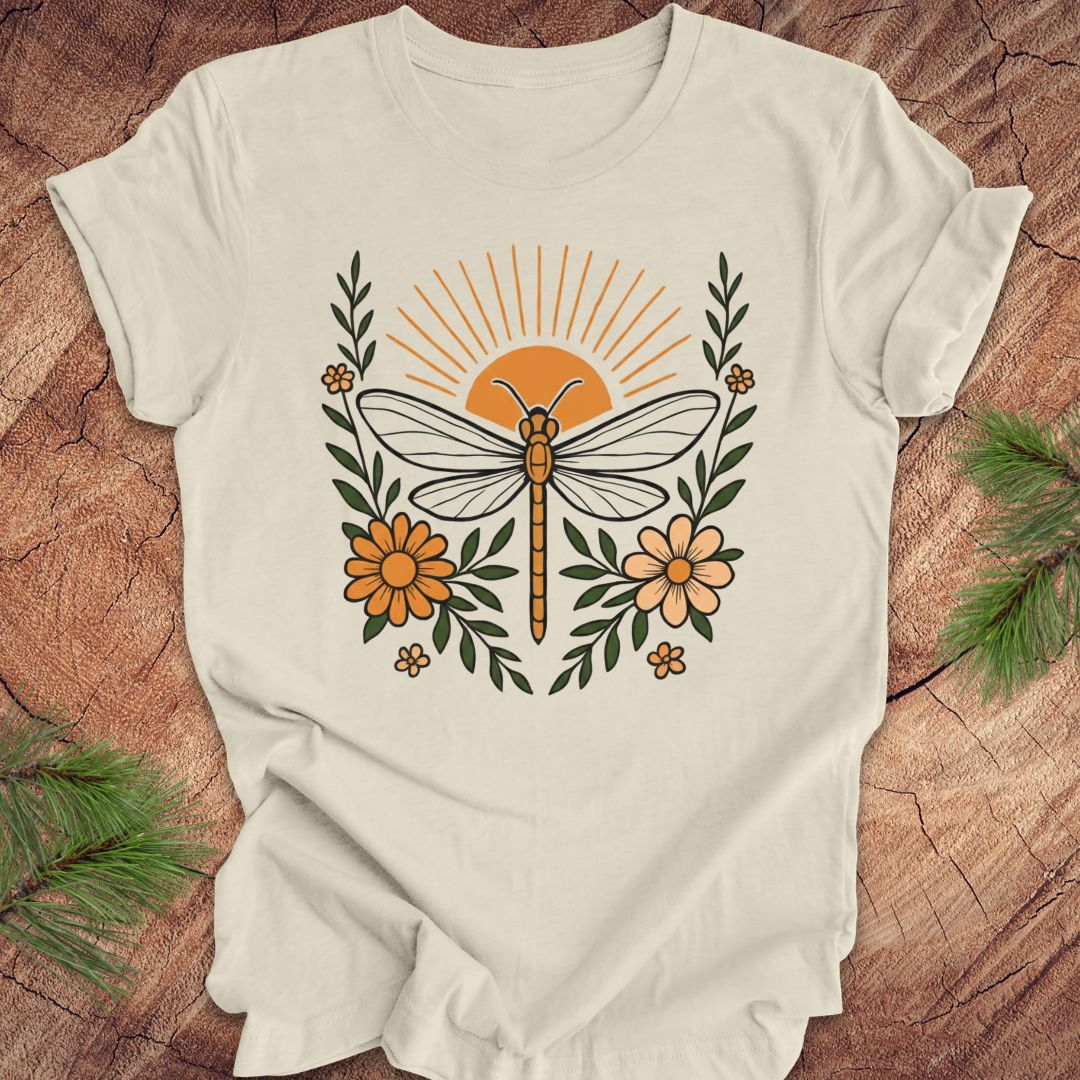 Beige t-shirt with a dragonfly, sunrise and flowers on a wood background.