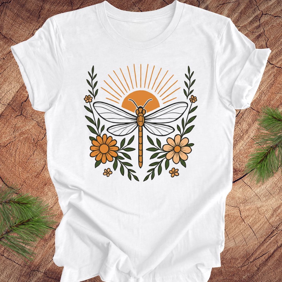 White t-shirt with a dragonfly, sunrise and flowers on a wood background.