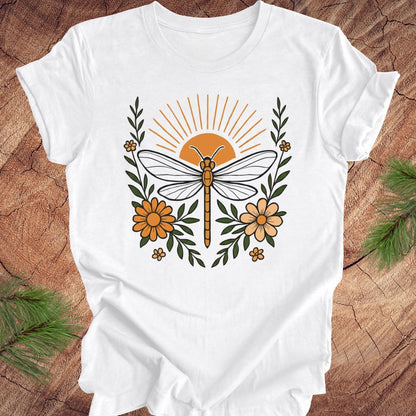 White t-shirt with a dragonfly, sunrise and flowers on a wood background.