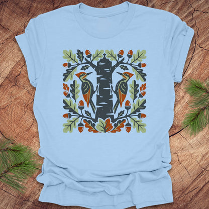 Light blue t-shirt with folk art woodpeckers in a tree design on a wooden surface