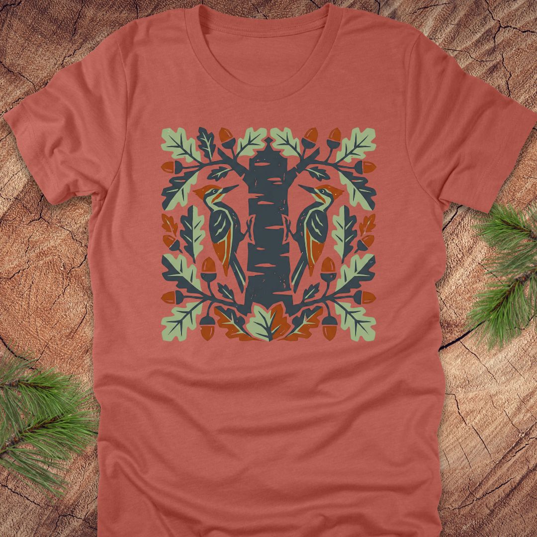 Clay colored t-shirt with folk art woodpeckers in a tree design on a wooden surface