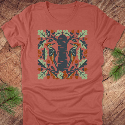 Clay colored t-shirt with folk art woodpeckers in a tree design on a wooden surface