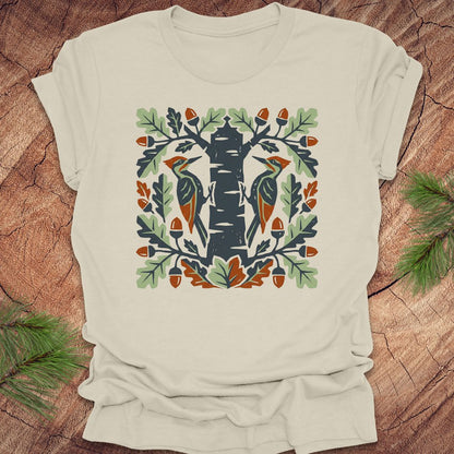 Beige t-shirt with folk art woodpeckers in a tree design on a wooden surface