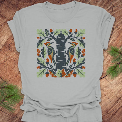 Light gray t-shirt with folk art woodpeckers in a tree design on a wooden surface