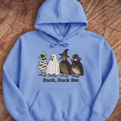 A light blue pullover hoodie laid flat on a textured wood background, featuring a graphic of four duck characters in Halloween costumes with the text 'Duck, Duck Boo' underneath.