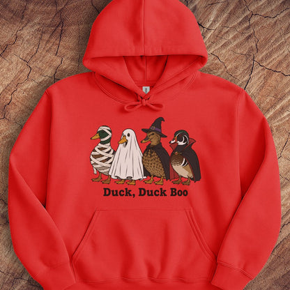 A bright red pullover hoodie laid flat on a textured wood background, featuring a front graphic of four cartoon ducks in Halloween costumes with the text 'Duck, Duck Boo' underneath.