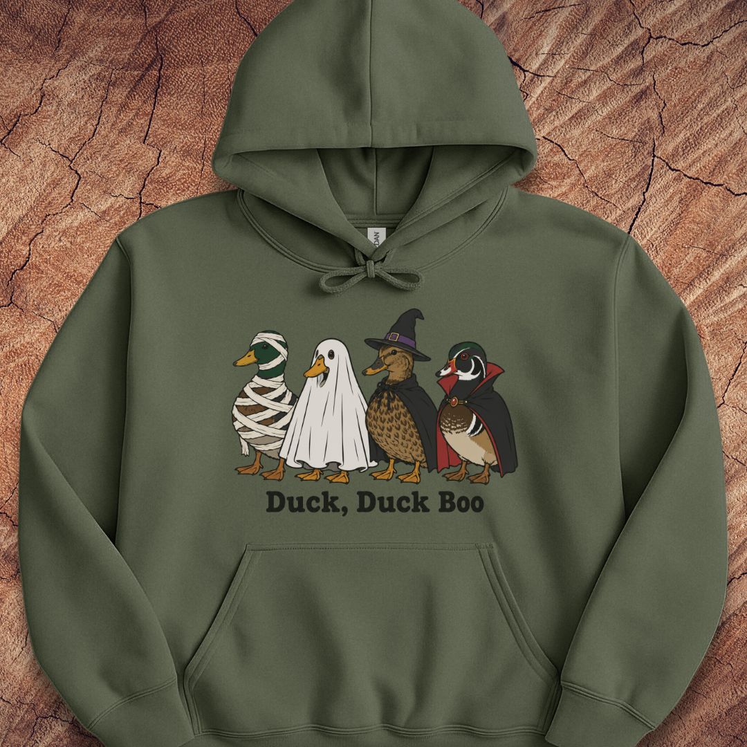 An olive green pullover hoodie featuring a graphic of four cartoon ducks in Halloween costumes (mummy, ghost, wizard, and woodcreeper) with the text 'Duck, Duck Boo' printed below. The hoodie is laid flat on a textured brown wood background.