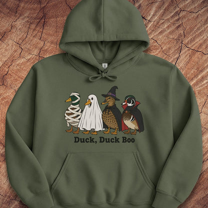An olive green pullover hoodie featuring a graphic of four cartoon ducks in Halloween costumes (mummy, ghost, wizard, and woodcreeper) with the text 'Duck, Duck Boo' printed below. The hoodie is laid flat on a textured brown wood background.