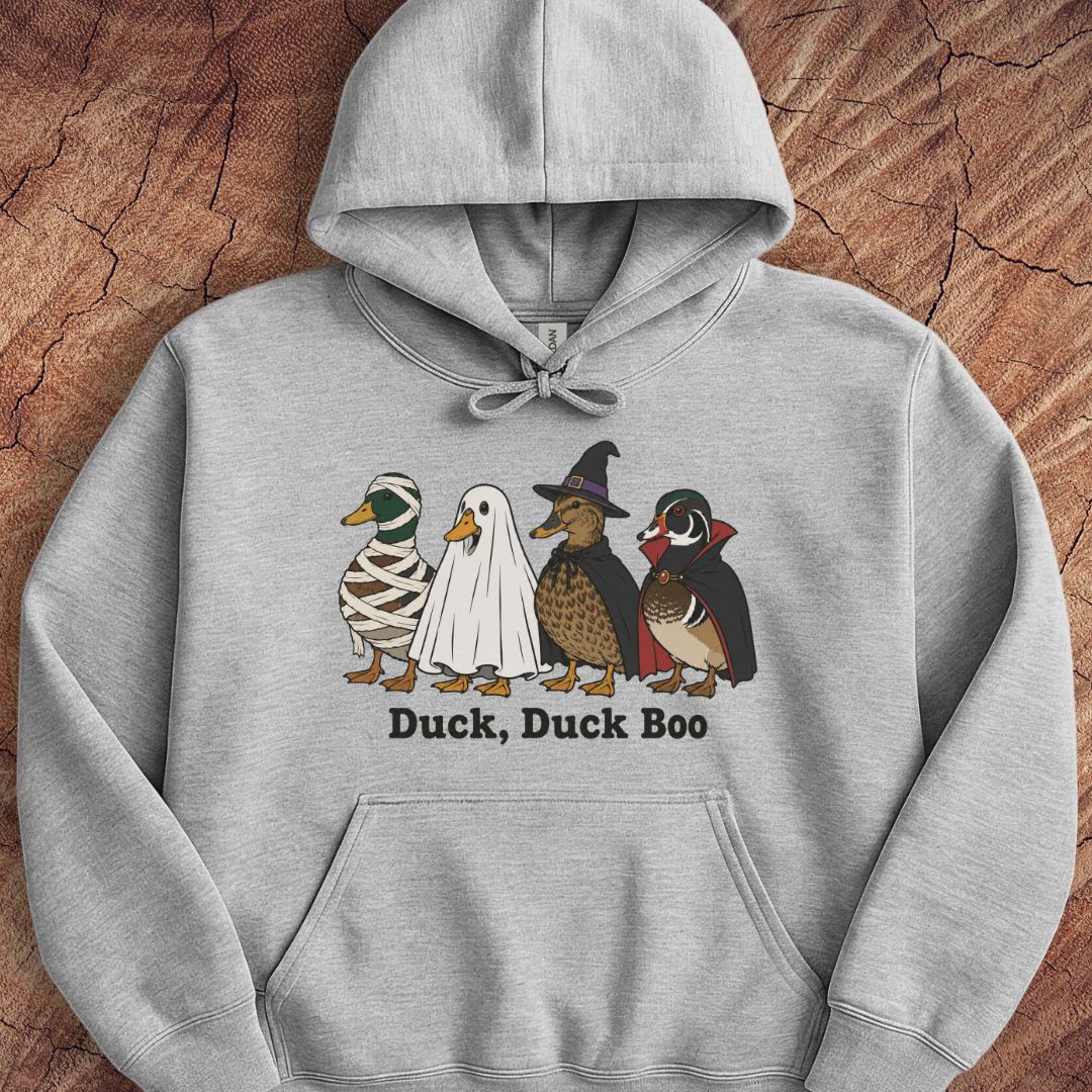 A light gray pullover hoodie laid flat on a textured brown wood background, featuring a graphic of four cartoon ducks dressed in Halloween costumes with the text 'Duck, Duck Boo' underneath.