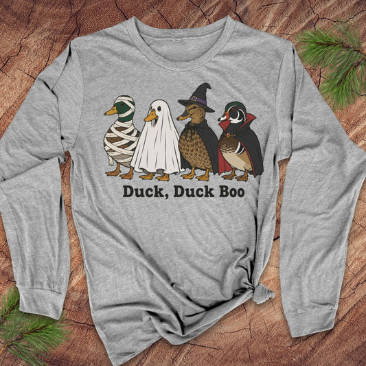 A heather gray long-sleeve t-shirt with a graphic of four ducks in Halloween costumes (mummy, ghost, witch, and Native American) above the text 'Duck, Duck Boo'. The shirt is displayed flat on a rustic wood background with pine needles.